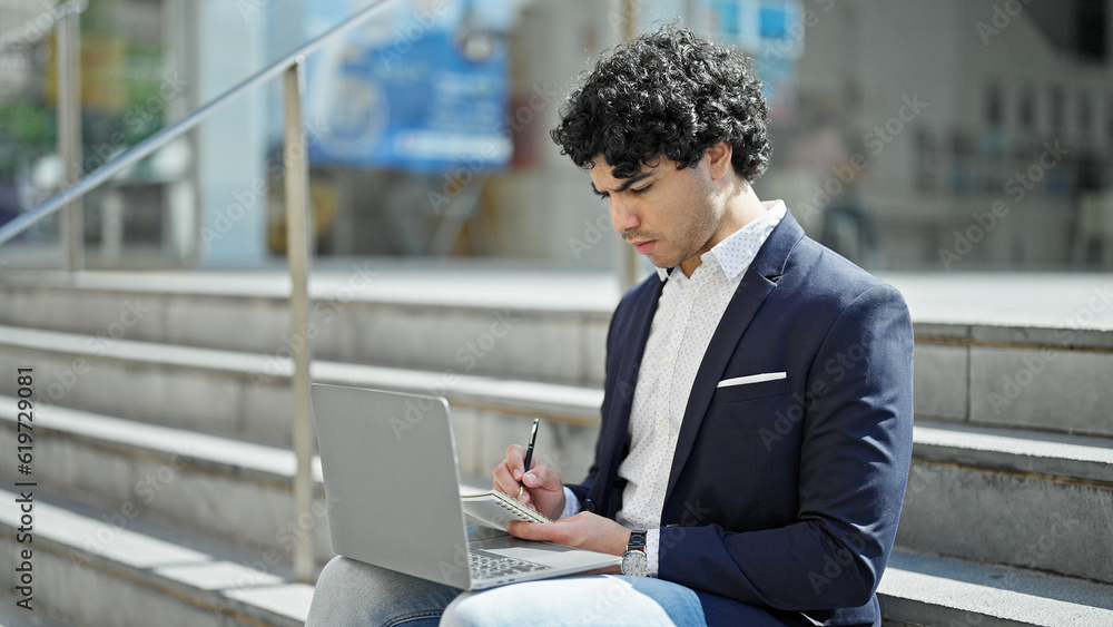 Young latin man business worker using laptop writing on notebook at street Young latin man business worker using laptop writing on notebook at street
