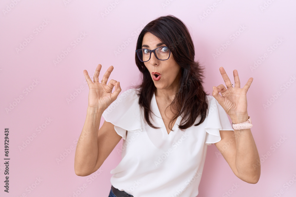 Middle age hispanic woman wearing casual white t shirt and glasses looking surprised and shocked doing ok approval symbol with fingers. crazy expression