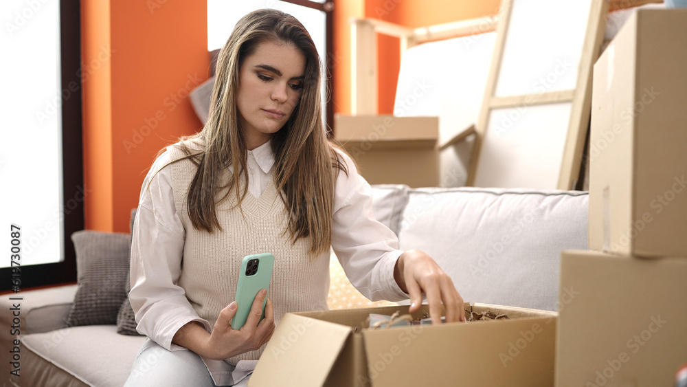 Young beautiful hispanic woman unpacking cardboard box using smartphone at new home Young beautiful hispanic woman unpacking cardboard box using smartphone at new home