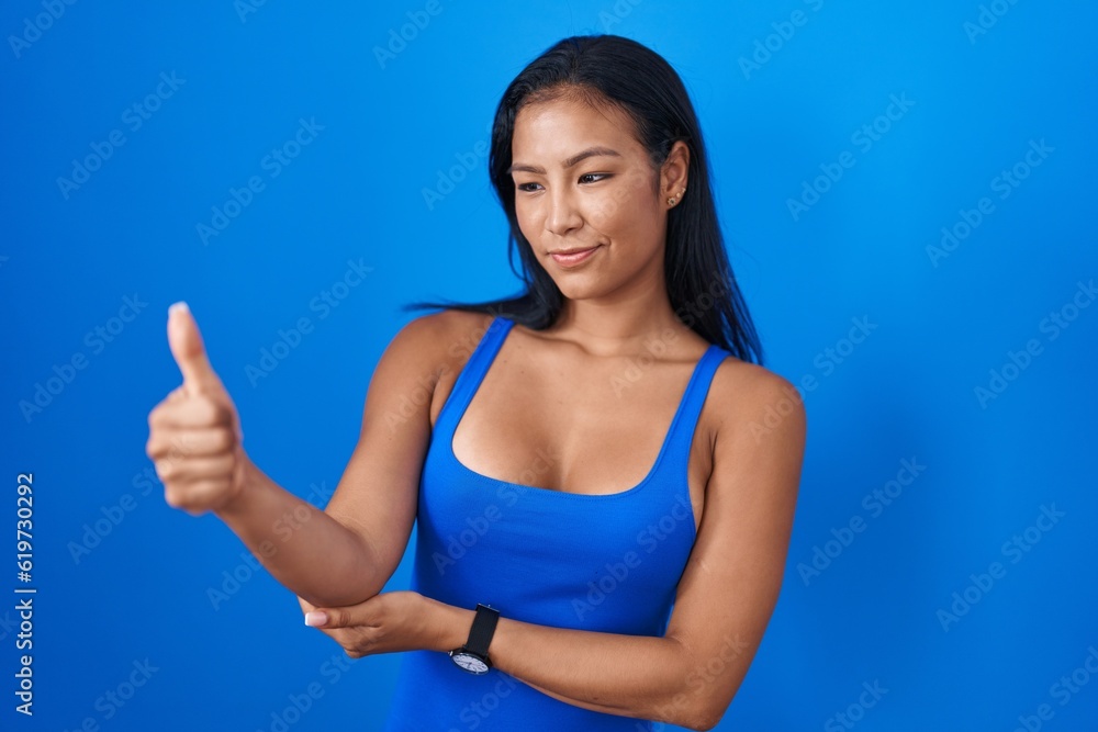 Hispanic woman standing over blue background looking proud, smiling doing thumbs up gesture to the side Hispanic woman standing over blue background looking proud, smiling doing thumbs up gesture to the side