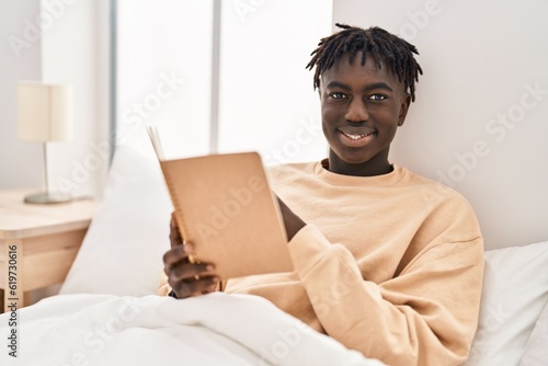 African american man reading book sitting on bed at bedroom