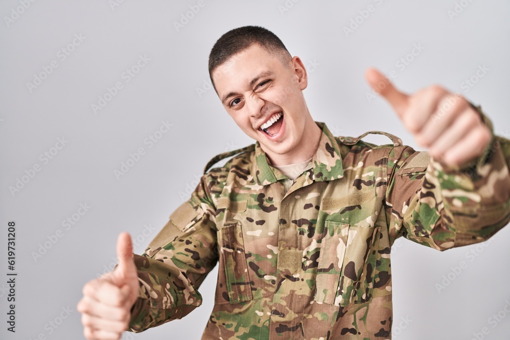 Young man wearing camouflage army uniform approving doing positive gesture with hand, thumbs up smiling and happy for success. winner gesture. Young man wearing camouflage army uniform approving doing positive gesture with hand, thumbs up smiling and happy for success. winner gesture.