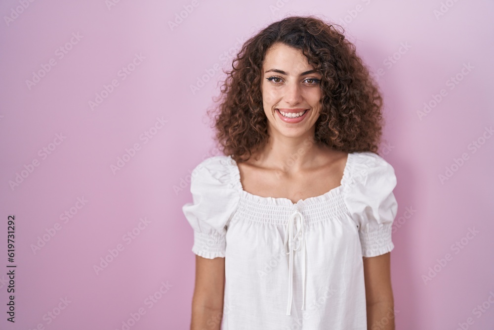 Hispanic woman with curly hair standing over pink background with a happy and cool smile on face. lucky person. Hispanic woman with curly hair standing over pink background with a happy and cool smile on face. lucky person.