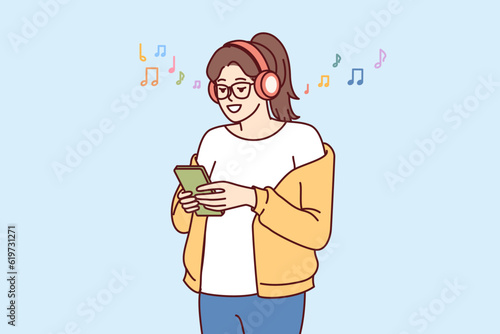 Teenage girl listens music on headphones and holds phone with application to select track from playlist. Young woman meloman uses wireless headphones and smartphone to search for favorite music