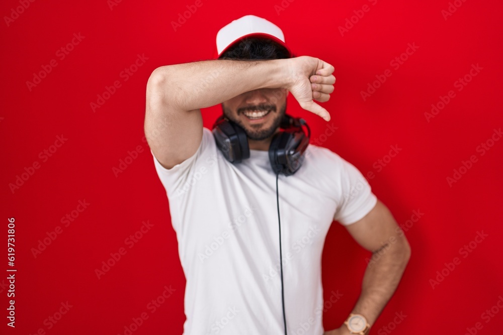 Hispanic man with beard wearing gamer hat and headphones covering eyes with arm smiling cheerful and funny. blind concept. Hispanic man with beard wearing gamer hat and headphones covering eyes with arm smiling cheerful and funny. blind concept.