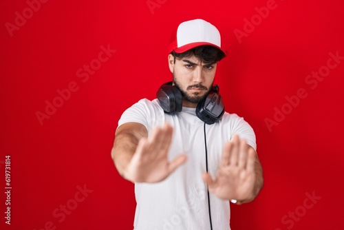 Hispanic man with beard wearing gamer hat and headphones moving away hands palms showing refusal and denial with afraid and disgusting expression. stop and forbidden.