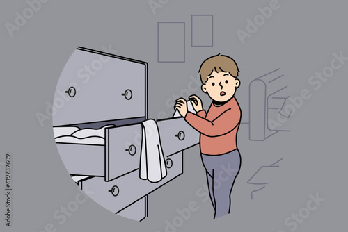 Little boy is standing near closet with clothes, looking at textiles with curiosity and studying world around. Childish pranks of funny boy throwing clothes from drawer making mess while playing