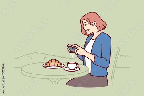 Woman blogger takes pictures of breakfast sitting in cafe and posting pictures of food on social network. Girl teenager blogger in street restaurant making photo of croissant and cup of coffee