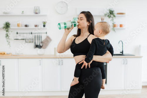 Healthy young lady in yoga clothes drinking water from sports bottle while keeping baby on left side in kitchen interior. Beautiful slim mother maintaining hydration before starting training at home.