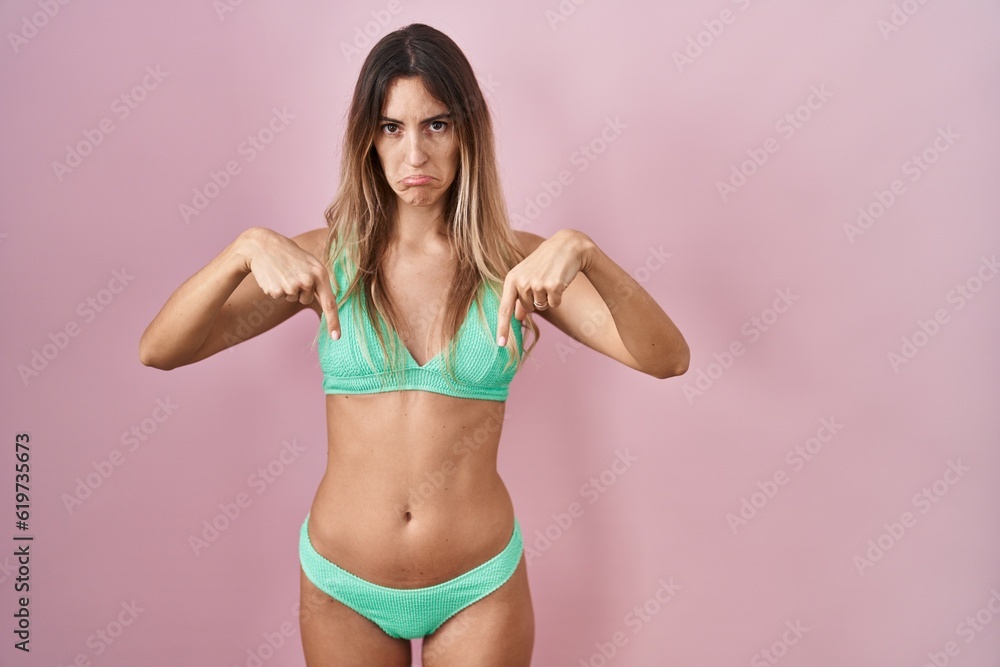 Naklejka premium Young hispanic woman wearing bikini over pink background pointing down looking sad and upset, indicating direction with fingers, unhappy and depressed.