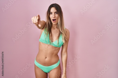 Young hispanic woman wearing bikini over pink background pointing with finger surprised ahead, open mouth amazed expression, something on the front © Krakenimages.com