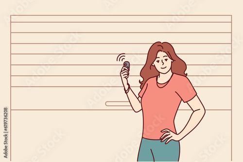 Woman stands near garage door pvc and uses electronic key to automatically open or close entrance. Girl driver poses at garage to securely store own car and protect personal vehicles from theft