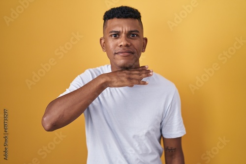 Young hispanic man standing over yellow background cutting throat with hand as knife, threaten aggression with furious violence