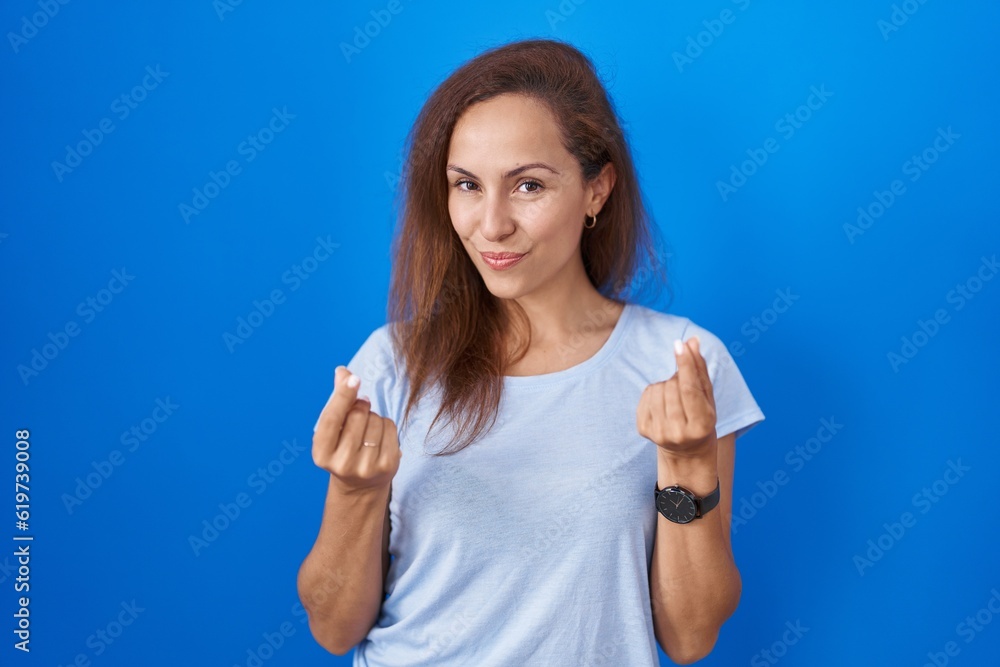 Fototapeta premium Brunette woman standing over blue background doing money gesture with hands, asking for salary payment, millionaire business