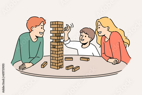 Happy family plays jenga with child enjoying board game and developing fine motor skills in baby. Satisfied boy plays jenga with parents pulling wooden block out of unstable tower.