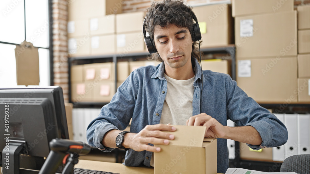 Young hispanic man ecommerce business worker using computer and headphones closing cardboard box at office Young hispanic man ecommerce business worker using computer and headphones closing cardboard box at office