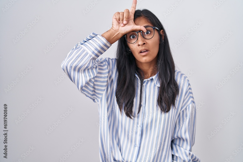 Naklejka premium Young hispanic woman wearing glasses making fun of people with fingers on forehead doing loser gesture mocking and insulting.