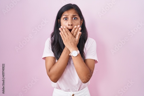 Young hispanic woman standing over pink background shocked covering mouth with hands for mistake. secret concept.