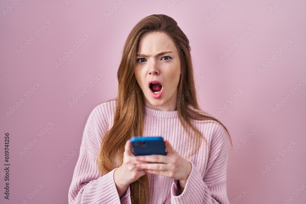 Young caucasian woman using smartphone typing message angry and mad screaming frustrated and furious, shouting with anger. rage and aggressive concept. Young caucasian woman using smartphone typing message angry and mad screaming frustrated and furious, shouting with anger. rage and aggressive concept.