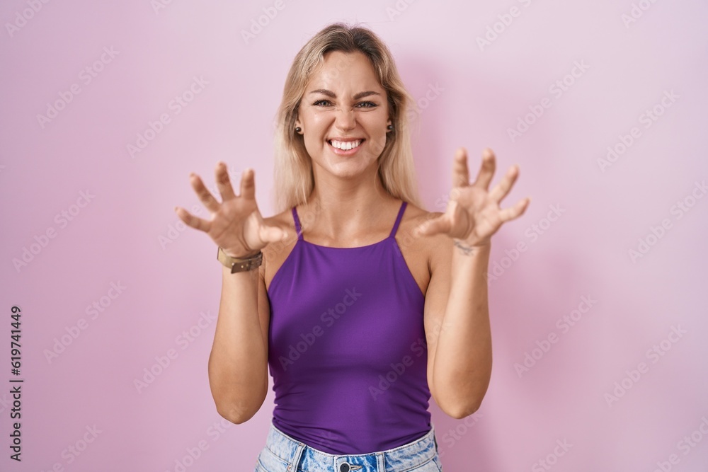 Young blonde woman standing over pink background smiling funny doing claw gesture as cat, aggressive and sexy expression Young blonde woman standing over pink background smiling funny doing claw gesture as cat, aggressive and sexy expression