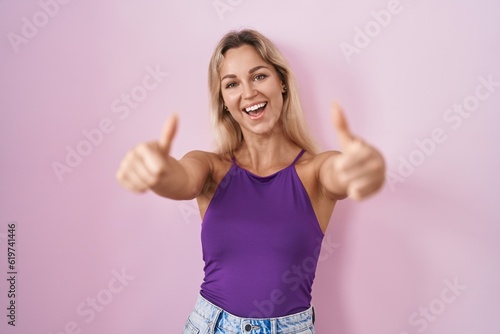 Young blonde woman standing over pink background approving doing positive gesture with hand  thumbs up smiling and happy for success. winner gesture.