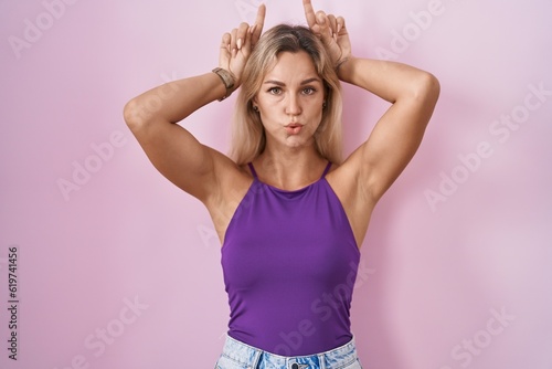 Young blonde woman standing over pink background doing funny gesture with finger over head as bull horns