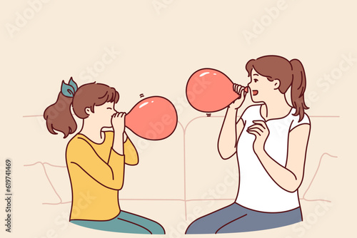 Mother and daughter inflate balloons sitting on sofa to decorate apartment for birthday party. Girl together with older sister, uses balloons to play or inhale helium to change voice