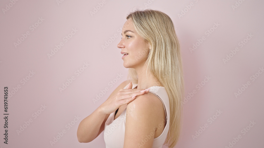 Young blonde woman applying skin treatment on shoulder over isolated pink background