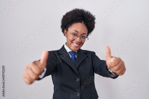 Beautiful african woman with curly hair wearing business jacket and glasses approving doing positive gesture with hand, thumbs up smiling and happy for success. winner gesture.