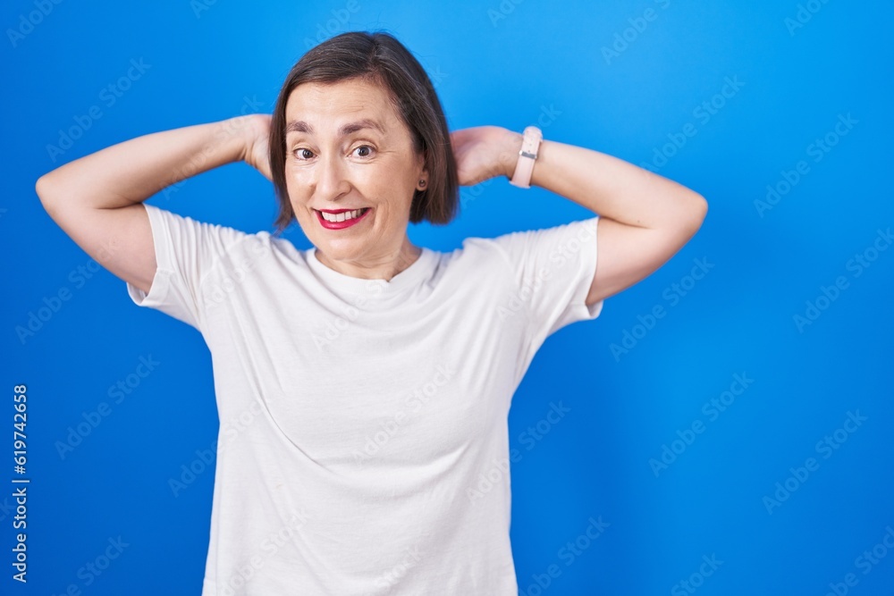 Fototapeta premium Middle age hispanic woman standing over blue background relaxing and stretching, arms and hands behind head and neck smiling happy