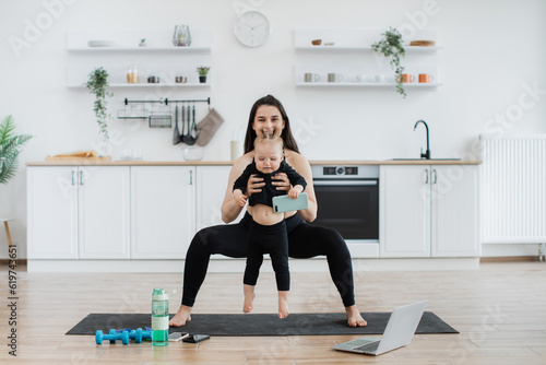 Attractive caucasian adult in athletic wear practising wide squats with baby in hands in kitchen interior. Active young mother engaging cute baby daughter to physical activities indoors on sunny day.
