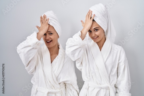 Middle age woman and daughter wearing white bathrobe and towel surprised with hand on head for mistake, remember error. forgot, bad memory concept.