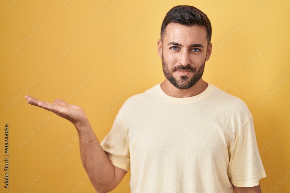 Handsome hispanic man standing over yellow background smiling cheerful presenting and pointing with palm of hand looking at the camera. Handsome hispanic man standing over yellow background smiling cheerful presenting and pointing with palm of hand looking at the camera.