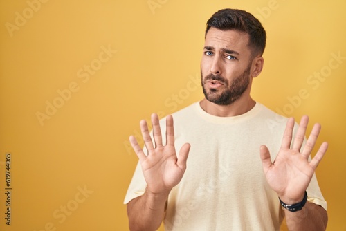 Handsome hispanic man standing over yellow background moving away hands palms showing refusal and denial with afraid and disgusting expression. stop and forbidden.