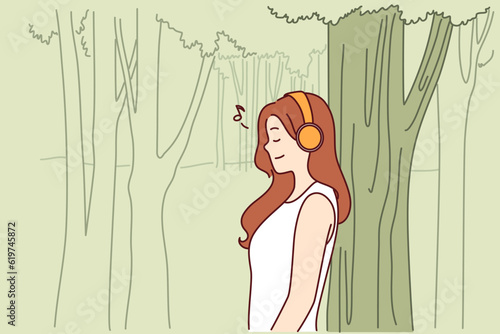 Woman listens to music through headphones standing in park near tall tree and enjoying good weather and fresh air. Teenager girl listens to music relaxing alone in nature after hard day at school