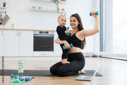 Athletic female in sports clothes embracing small kid while posing with weights on yoga mat inside house. Delighted family woman increasing body wellness while promoting active lifestyle indoors.