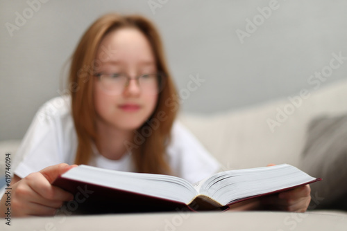 The girl is reading a thick book lying on the sofa.