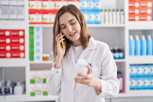 Young woman pharmacist talking on smartphone holding pills at pharmacy