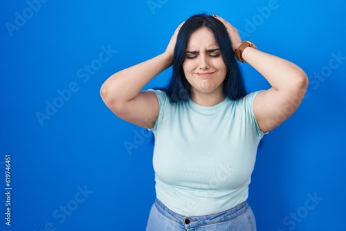 Young modern girl with blue hair standing over blue background suffering from headache desperate and stressed because pain and migraine. hands on head.