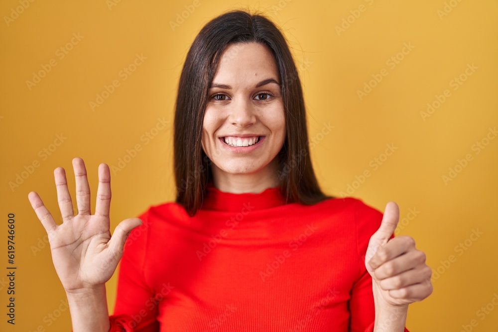 Young hispanic woman standing over yellow background showing and pointing up with fingers number six while smiling confident and happy. Young hispanic woman standing over yellow background showing and pointing up with fingers number six while smiling confident and happy.