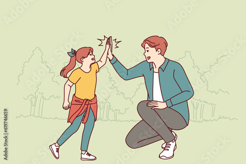 Dad gives hi-five to little daughter during walk together in park in hot summer weather. Cheerful teenager girl together with young dad is resting in nature enjoying active rest on sunday