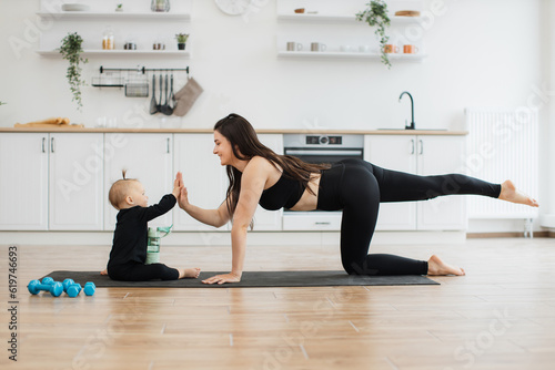 Funny small kid in black outfit responding to lady's high-five gesture while sitting near parent on floor. Affectionate mother establishing strong connection between her and child during home workout.