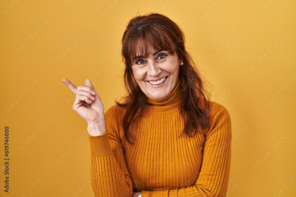 Middle age hispanic woman standing over yellow background with a big smile on face, pointing with hand finger to the side looking at the camera. Middle age hispanic woman standing over yellow background with a big smile on face, pointing with hand finger to the side looking at the camera.