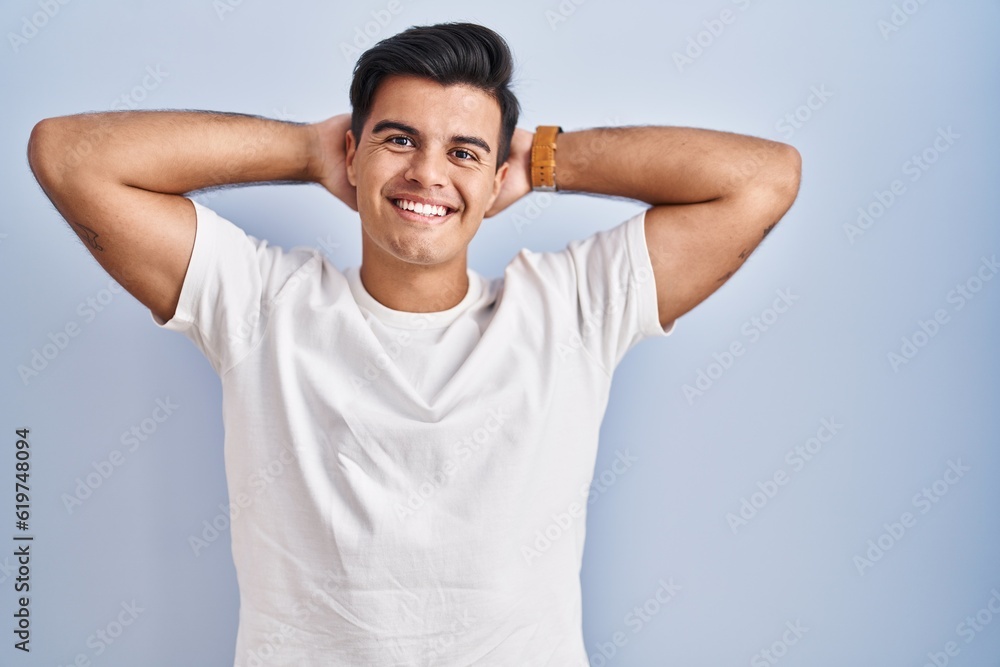 Hispanic man standing over blue background relaxing and stretching, arms and hands behind head and neck smiling happy Hispanic man standing over blue background relaxing and stretching, arms and hands behind head and neck smiling happy