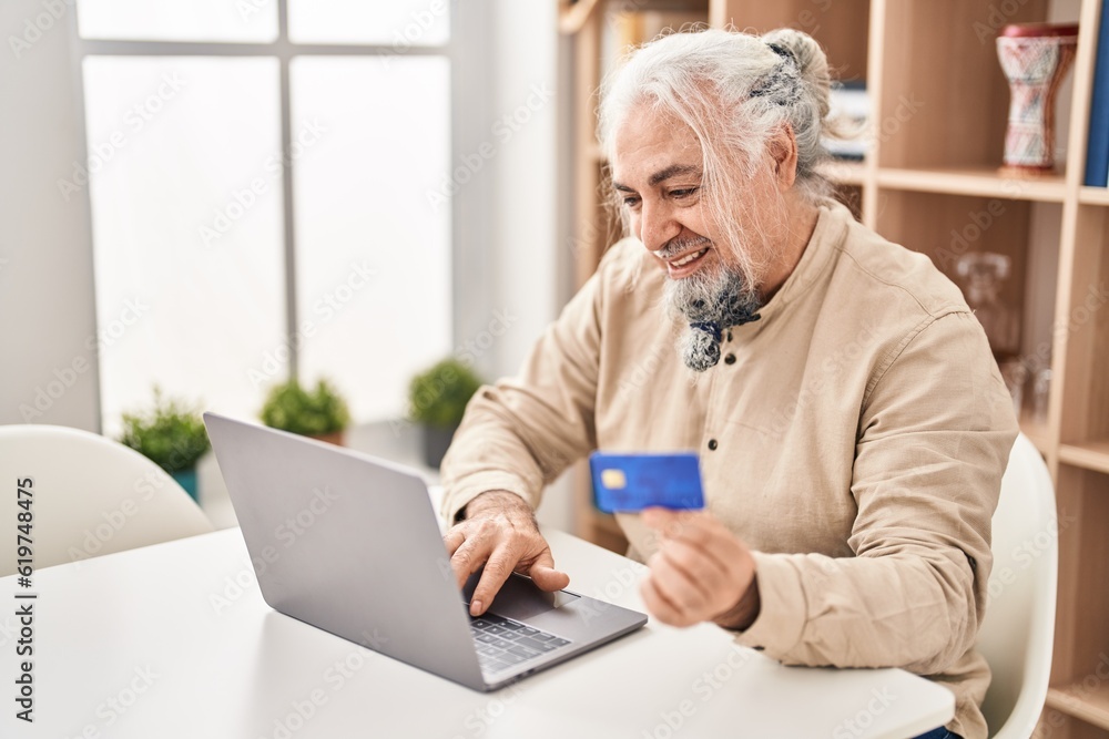 Middle age grey-haired man using laptop and credit card sitting on table at home Middle age grey-haired man using laptop and credit card sitting on table at home