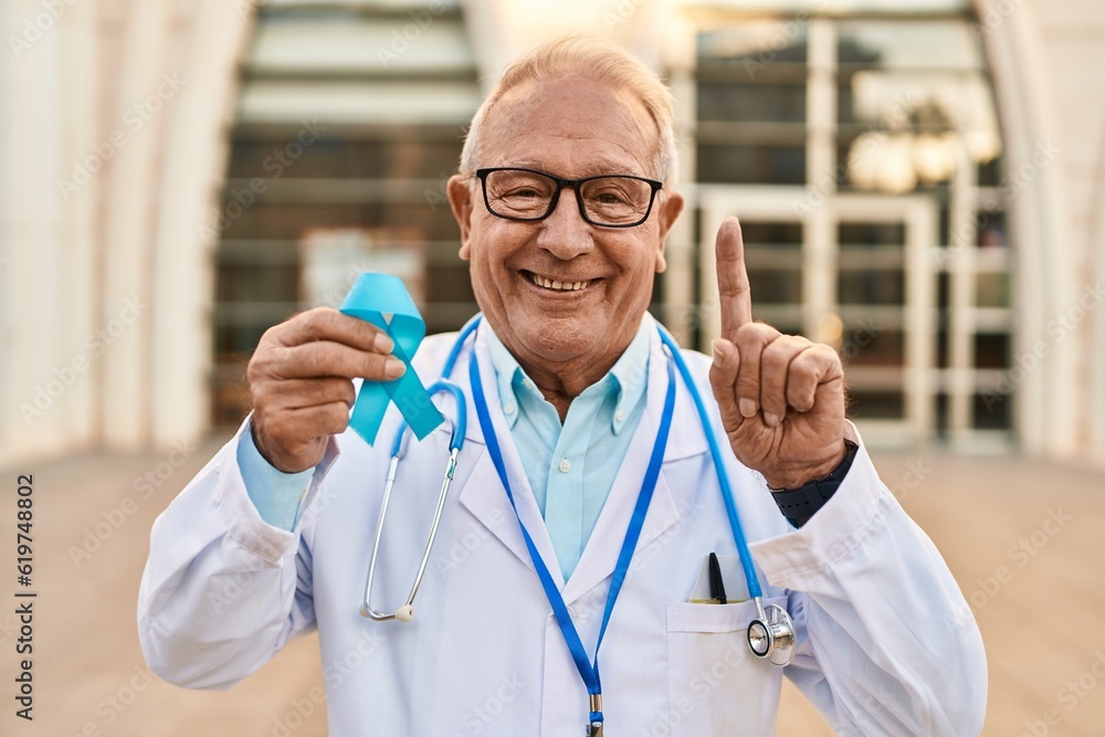 Senior doctor with grey hair holding blue ribbon surprised with an idea or question pointing finger with happy face, number one Senior doctor with grey hair holding blue ribbon surprised with an idea or question pointing finger with happy face, number one