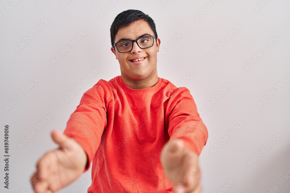 Young hispanic man with down syndrome standing over white background looking at the camera smiling with open arms for hug. cheerful expression embracing happiness. Young hispanic man with down syndrome standing over white background looking at the camera smiling with open arms for hug. cheerful expression embracing happiness.
