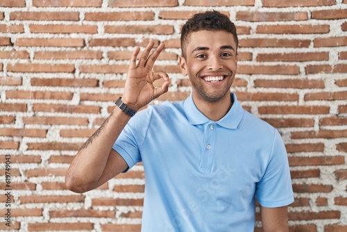 Brazilian young man standing over brick wall smiling positive doing ok sign with hand and fingers. successful expression.