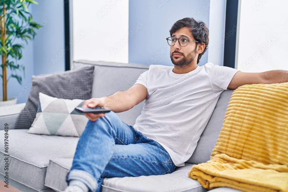 Handsome latin man holding television remote control smiling looking to the side and staring away thinking. Handsome latin man holding television remote control smiling looking to the side and staring away thinking.