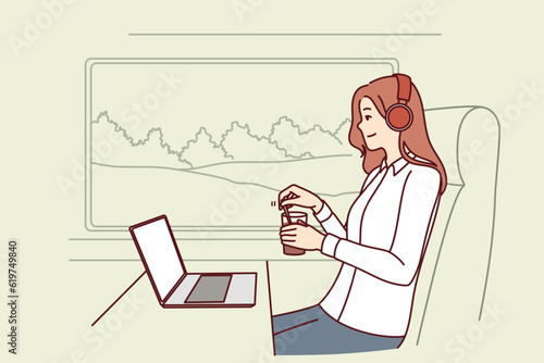 Woman train passenger uses laptop working on trip and drinks coffee sitting near large window in train carriage. Successful girl traveler in headphones sits in train watching movies